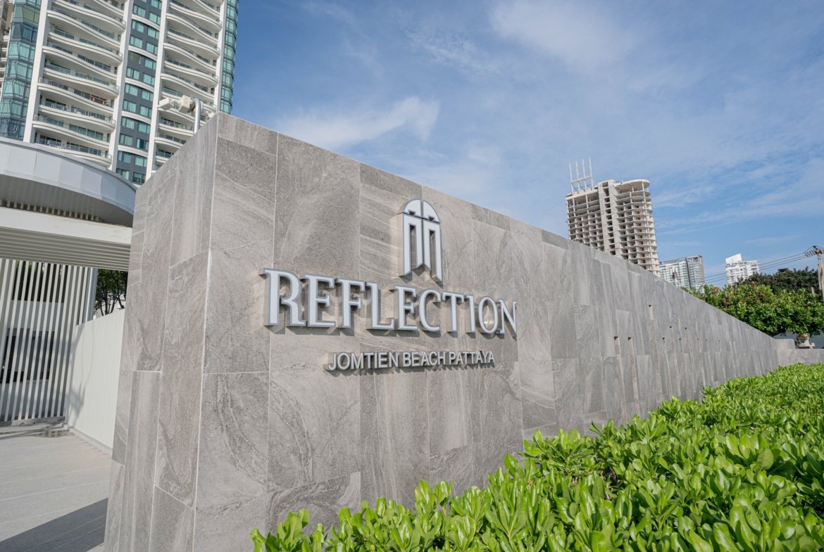 Stone wall sign reading 'REFLECTION' with 'Jomtien Beach Pattaya' below, set against modern high-rise buildings and a blue sky backdrop.