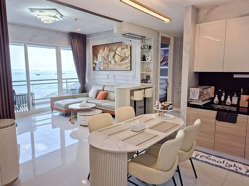 Bright, modern open-plan living space with large glass doors showing an ocean view, white furniture and marble surfaces throughout the lounge and dining area.