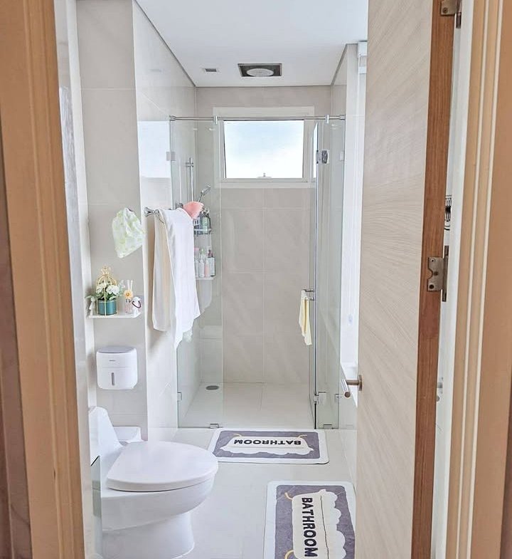 Bright modern bathroom: glass-walled shower, white tiles, towels on a rack, and two 'BATHROOM' mats on the floor.