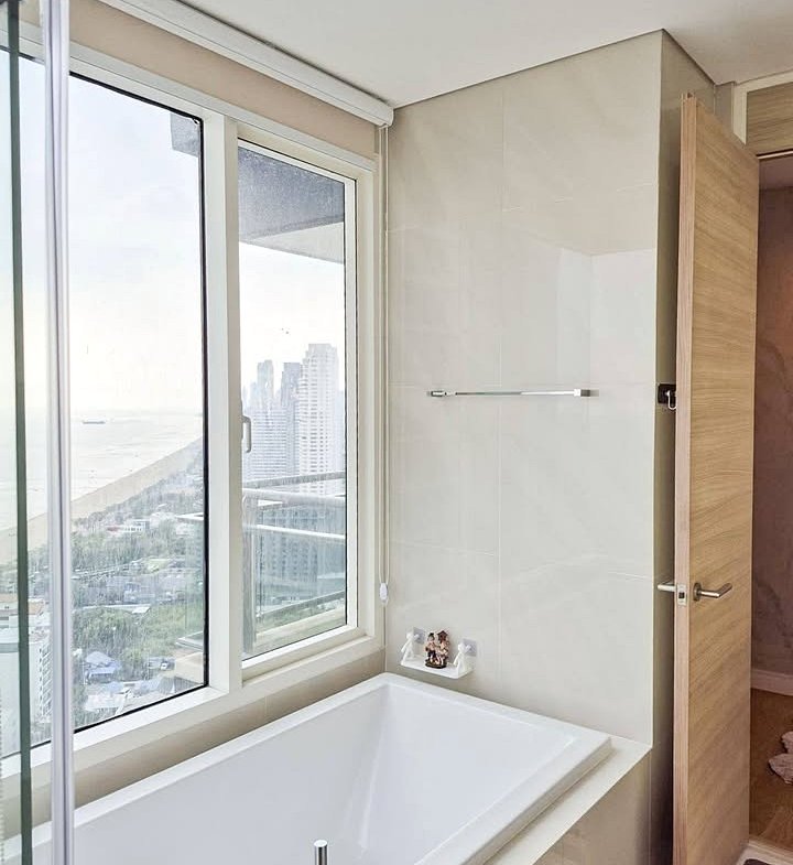 White rectangular bathtub against beige tiled wall beneath large windows with a city/ocean view outside, glass shower door on the left.