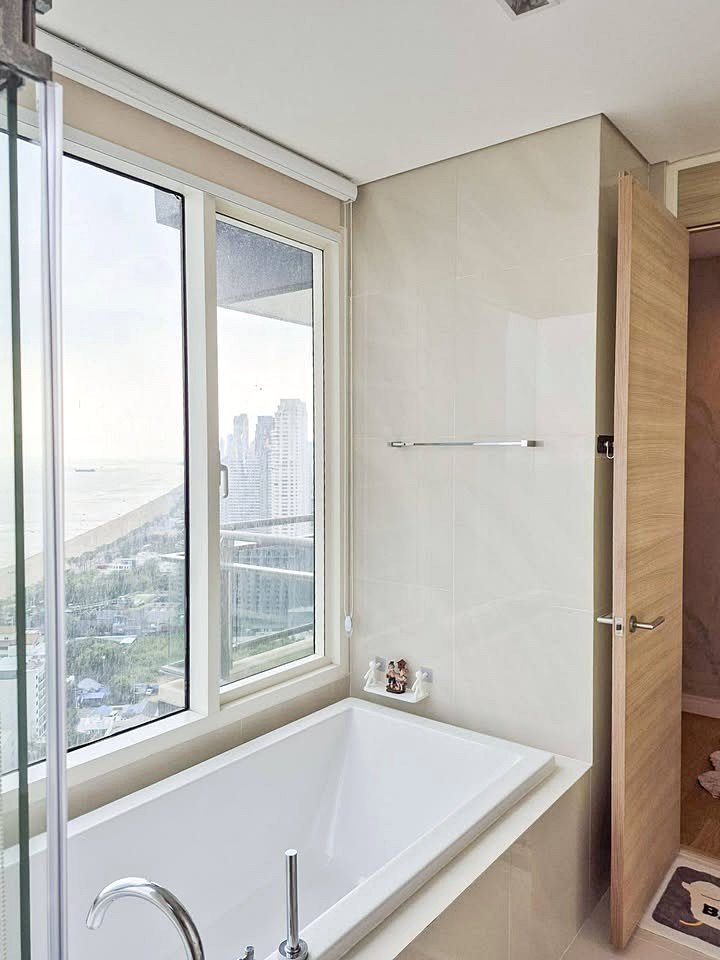 Reflection_Jomtien_Sale_Rent_Dlc242_11 White rectangular bathtub against beige tiled wall beneath large windows with a city/ocean view outside, glass shower door on the left.