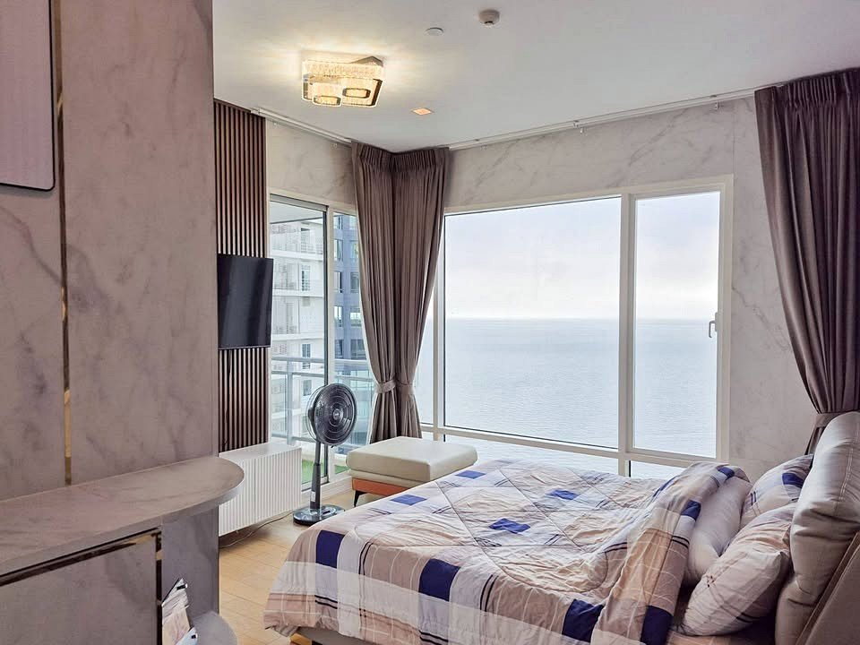 Bright hotel bedroom with a king bed facing a large bay window showing the ocean view; quilted blue-white bedding and pillows, dark curtains, and a fan nearby.