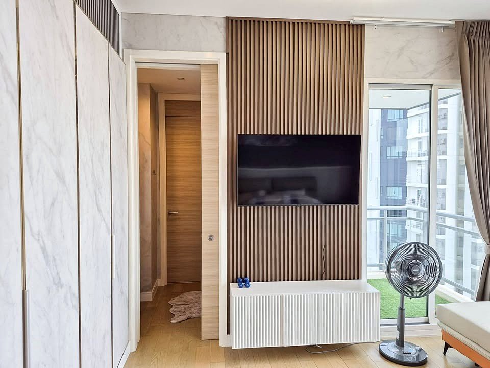 Living room with a wall-mounted flat-screen TV on a vertical wood slat feature, white radiator cabinet below, and a standing fan near a large window with a balcony view.