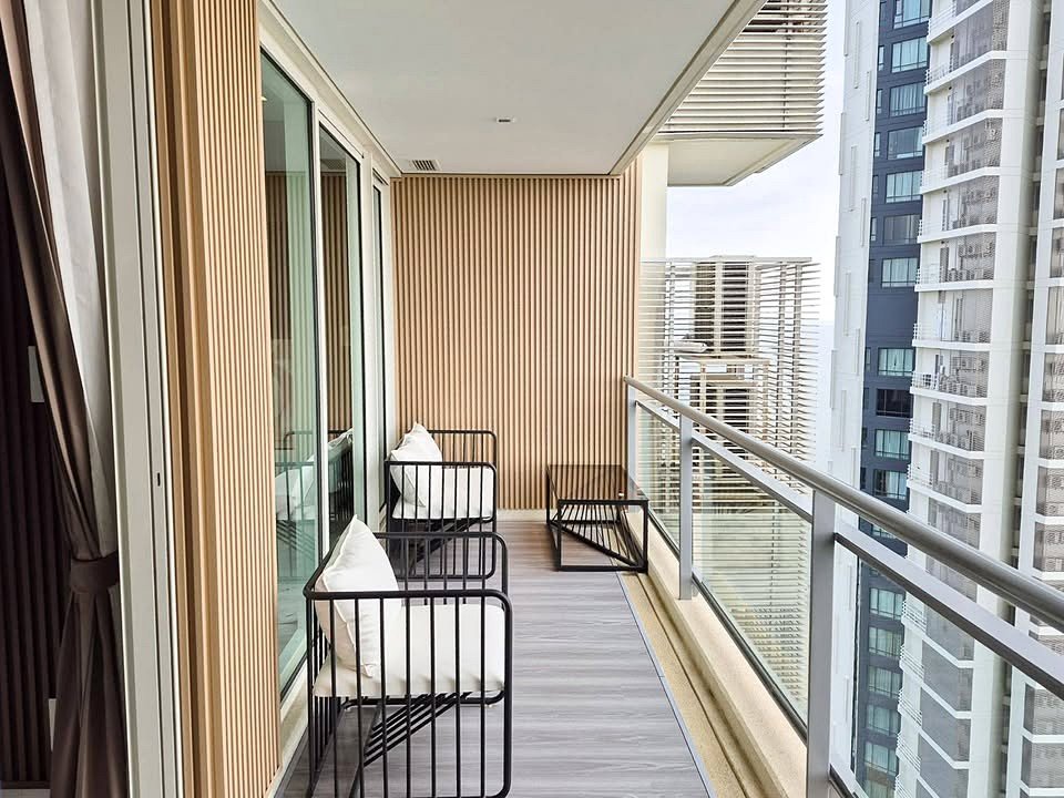 Modern apartment balcony with black metal chairs and white cushions, wooden slat wall, glass railing, overlooking neighboring high-rise buildings.