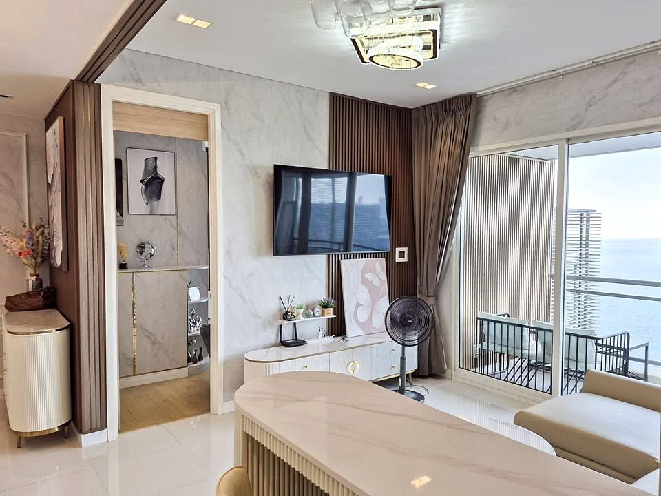 Bright contemporary living room with marble walls, a wall-mounted TV, and a balcony overlooking the ocean.