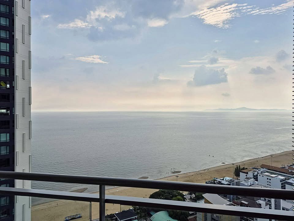 Balcony railing with a city beach and calm sea beyond, under a partly cloudy sky at sunset.