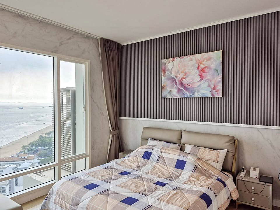 Bedroom with a large window showing an ocean view, a beige upholstered bed, and a dark vertical-grooved accent wall topped by a floral painting.