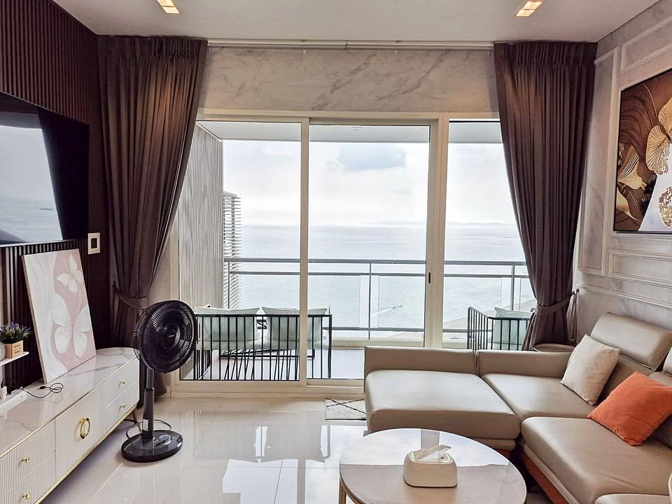 Modern living room with an L-shaped beige sofa and orange cushion facing a glass sliding door that opens to a balcony overlooking the sea.