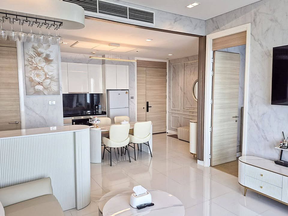 Open-concept living space with a curved white bar, six cream dining chairs, marble walls, and a modern kitchen in the background.