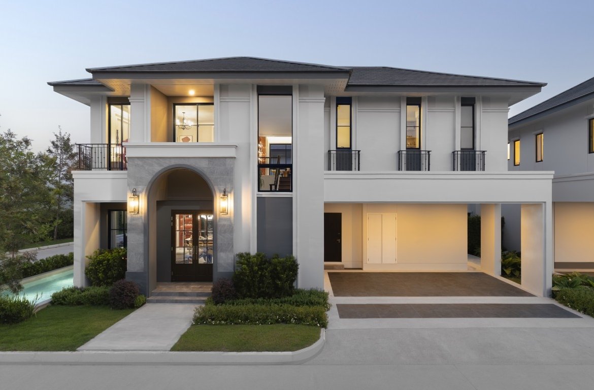 Front view of a modern two-story house with an arched entry, illuminated doorway, and a long driveway leading to a garage; pool and landscaping on the left.