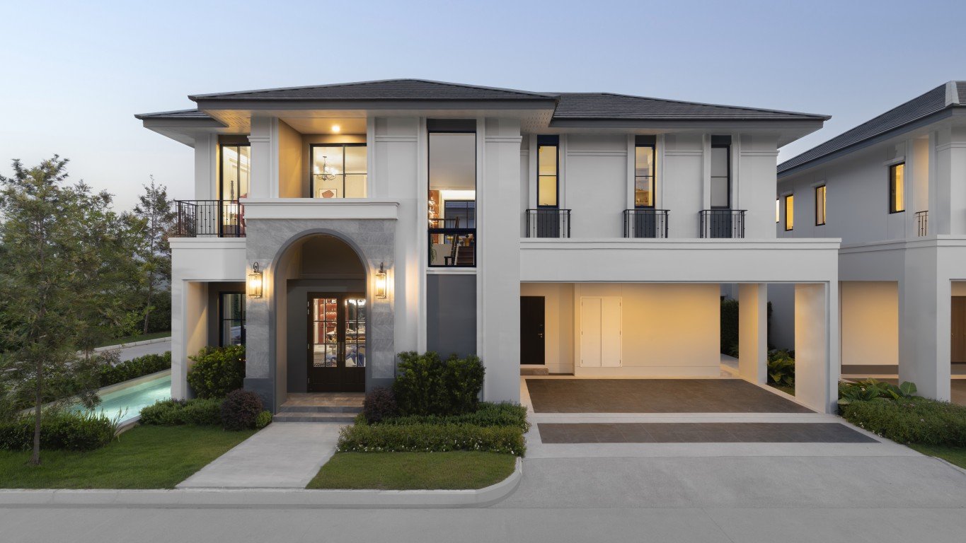 Rekha Pattaya-Motorway 15 Two-story modern house with a prominent arched entry, white-and-gray facade, and warm exterior lighting; driveway and pool on the left side