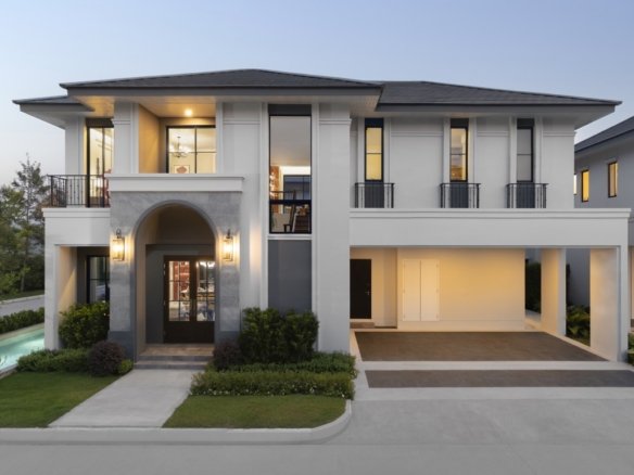 Front view of a modern two story white house with an arched entry black window frames and a driveway with a covered parking area pool visible to the left at dusk