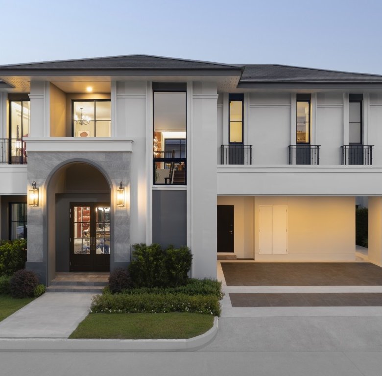 Front view of a modern two story white house with an arched entry black window frames and a driveway with a covered parking area pool visible to the left at dusk