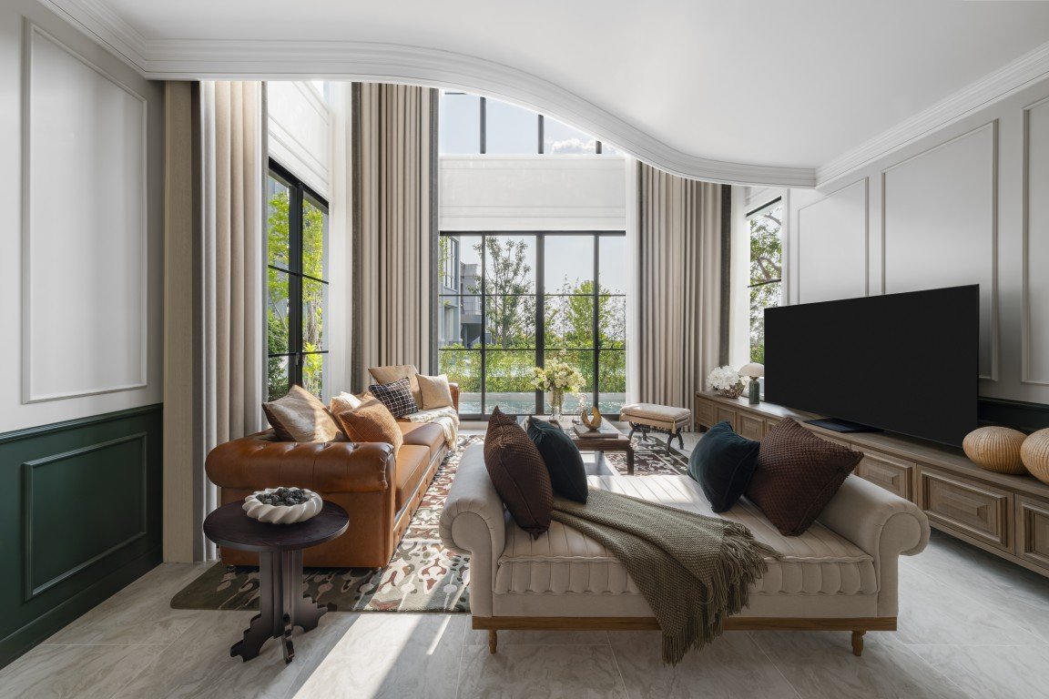 Sunlit living room with beige sofa, brown leather couch, large windows, and a flat-screen TV on wood cabinets.