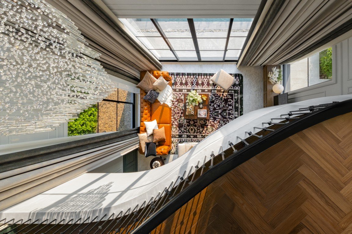 Top-down view of a modern living room seen from a curved staircase: orange sofa, patterned rug, coffee table, chandelier, and large windows in natural light.