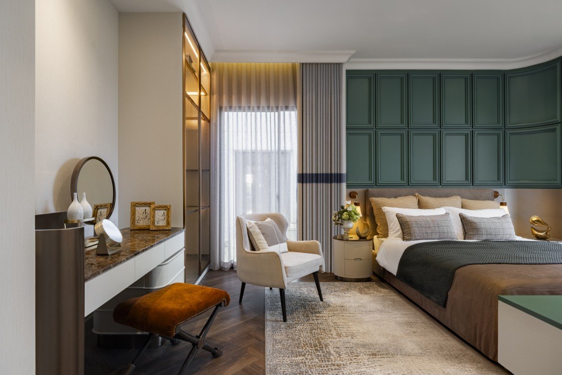 Luxurious hotel bedroom with a green paneled wall, king bed, and plush pillows; a seating chair, vanity desk, and mirror on the left.