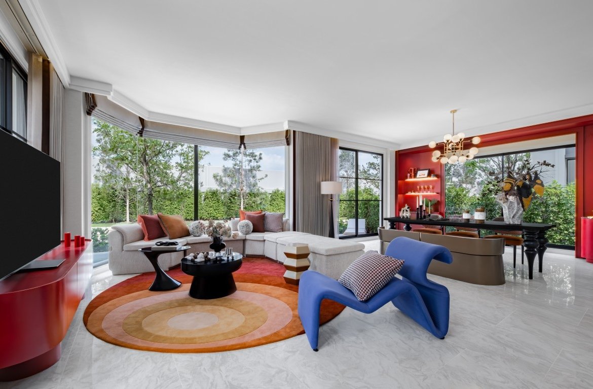 Modern living room with a white sectional sofa, circular orange rug, and large windows overlooking greenery outside.