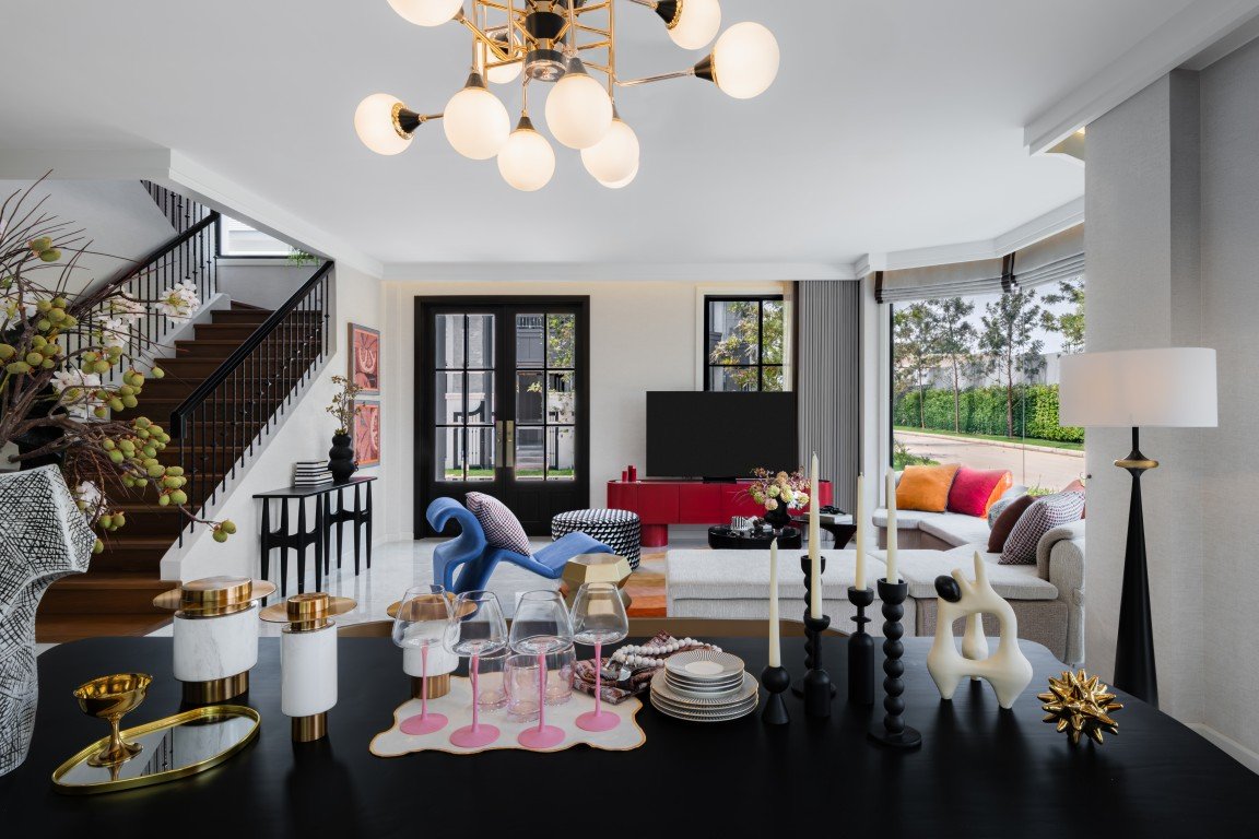 Open-concept living and dining area with a black table set with glassware and candles, a staircase on the left, and a sunlit seating area beyond.