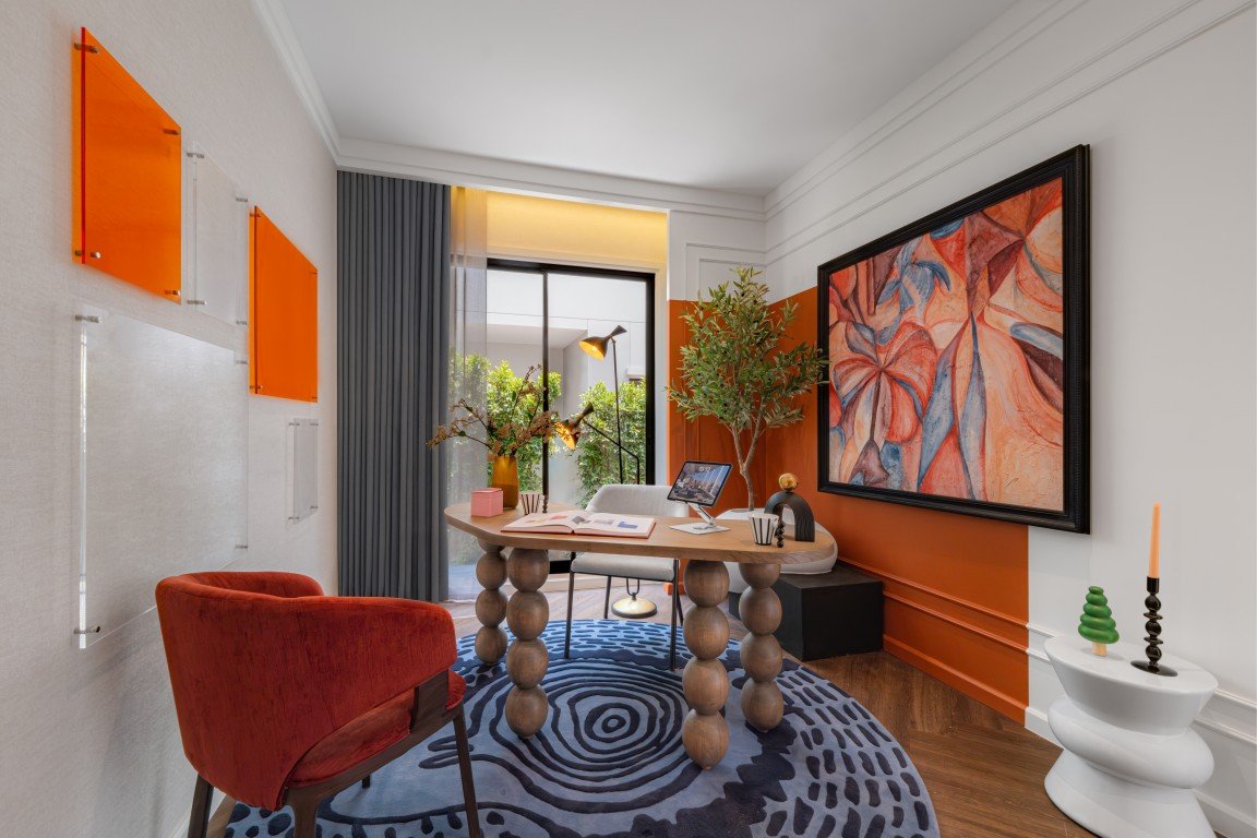 Contemporary home office with a round wooden table on a blue swirl rug, red chair, orange wall panels, and a large abstract painting.