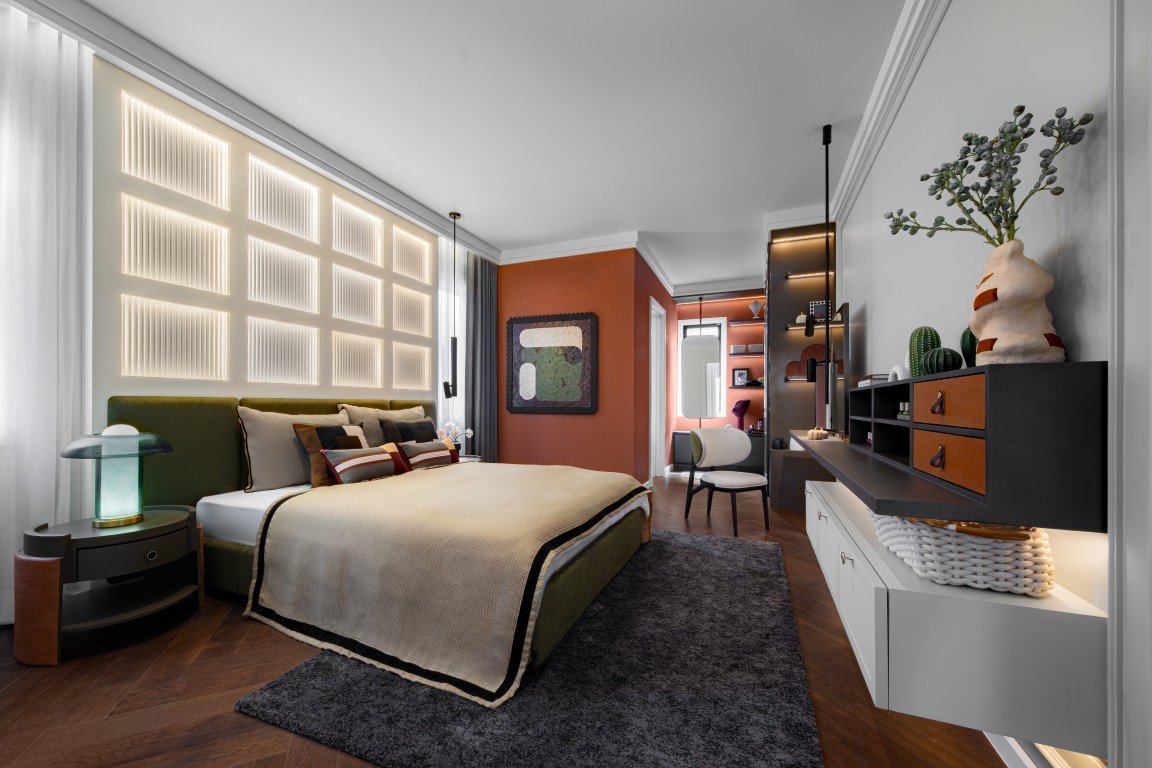 Modern hotel bedroom with an olive-green bed, orange accent wall, and a desk area on the right; warm, contemporary design and plush rug.