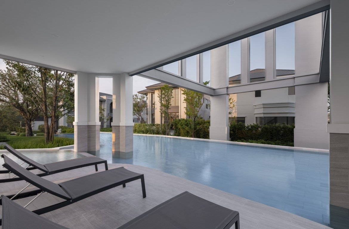 Poolside view of a modern covered terrace with lounge chairs beside a long blue pool and white columns.