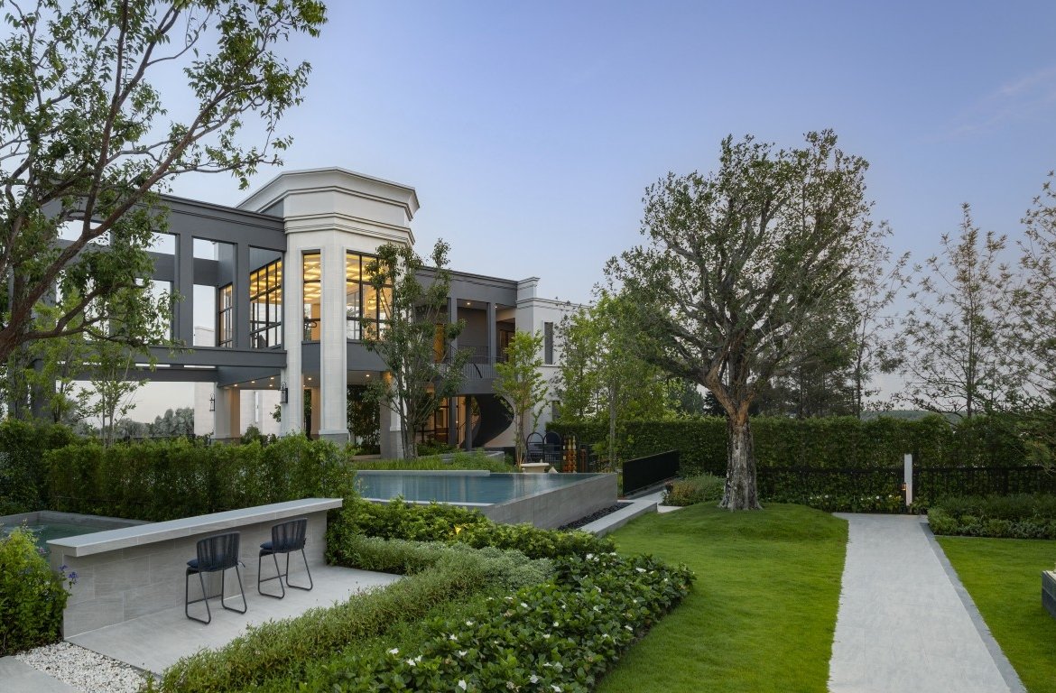 Modern two-story house with glass walls, a rectangular pool, and a manicured lawn with seating area.