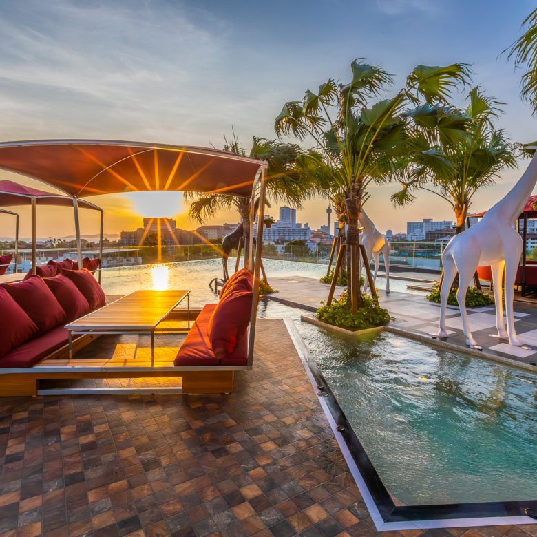 Sunset over a luxury rooftop lounge with red cushioned seating palm trees and white horse statues by a water channel Logo bottom right Riviera Group