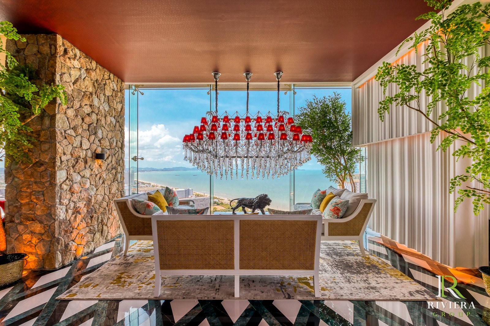 Riviera-Ocean-Drive-Pattaya 14 Luxurious living room with a red chandelier, stone wall, and floor-to-ceiling ocean views surrounded by greenery.