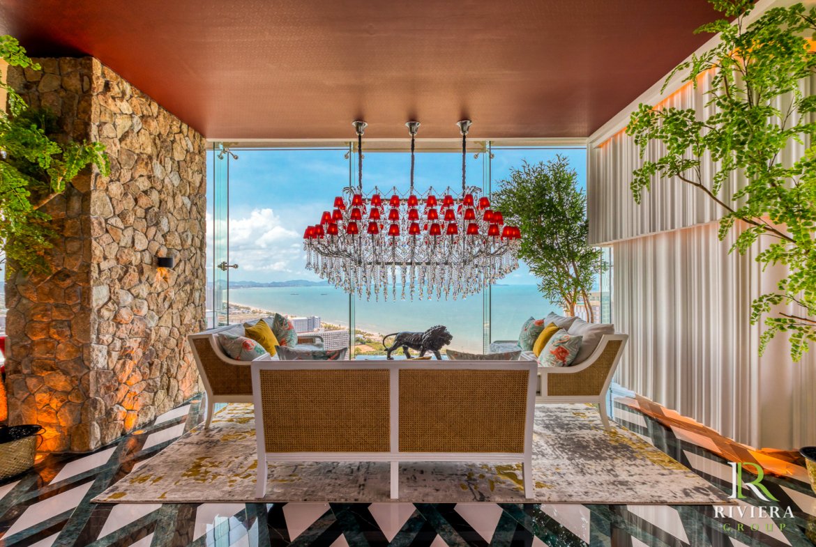 Luxurious living area with a cascading crystal chandelier of red lampshades, stone wall on the left, glass walls showing an ocean view, and wicker seating with colorful cushions. Green plants frame the space; Riviera Group logo in the bottom right.