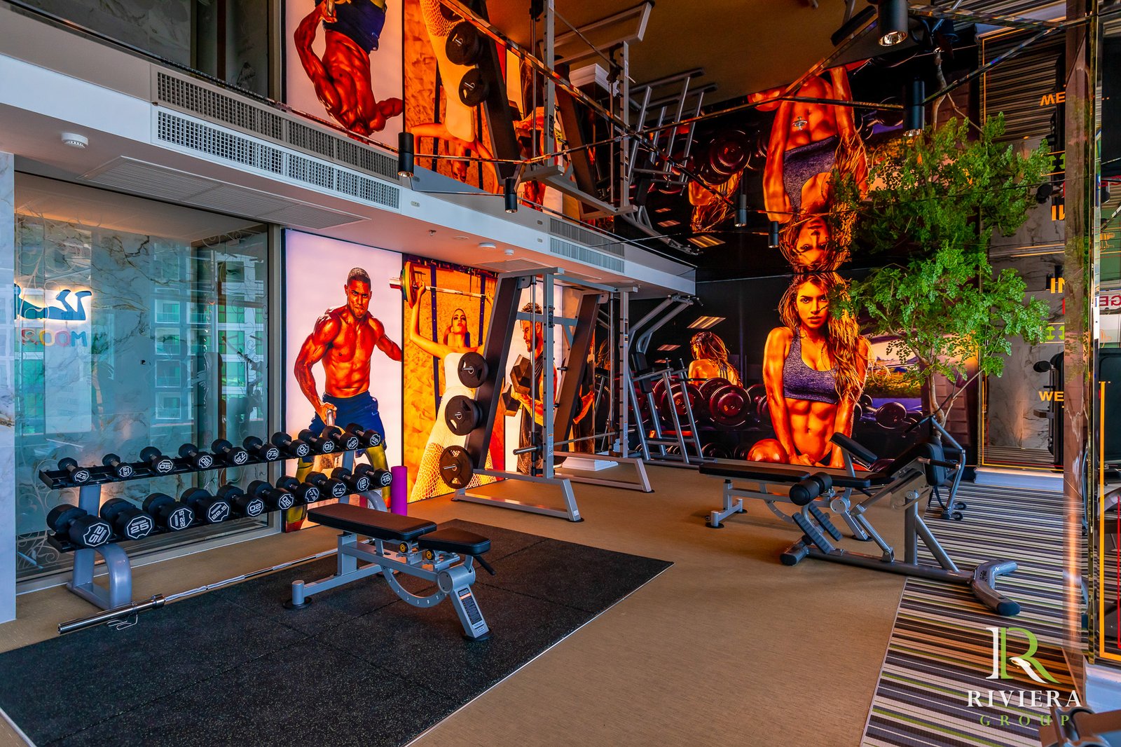 Riviera-Ocean-Drive-Pattaya 25 Gym interior with weight racks, benches, and colorful murals of muscular athletes on the walls; glass storefronts on the left.