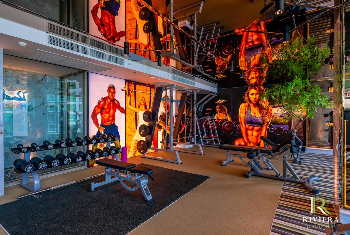 Gym interior with free weights, benches, and weight machines set against large wall murals of muscular athletes; glass storefront on the left and a striped entry mat along the floor.