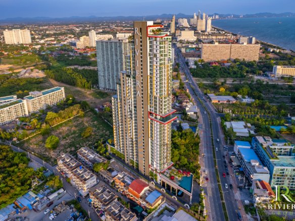 Aerial view of a coastal city with a tall modern residential tower labeled Riviera, surrounded by buildings, streets, and the ocean in the distance.
