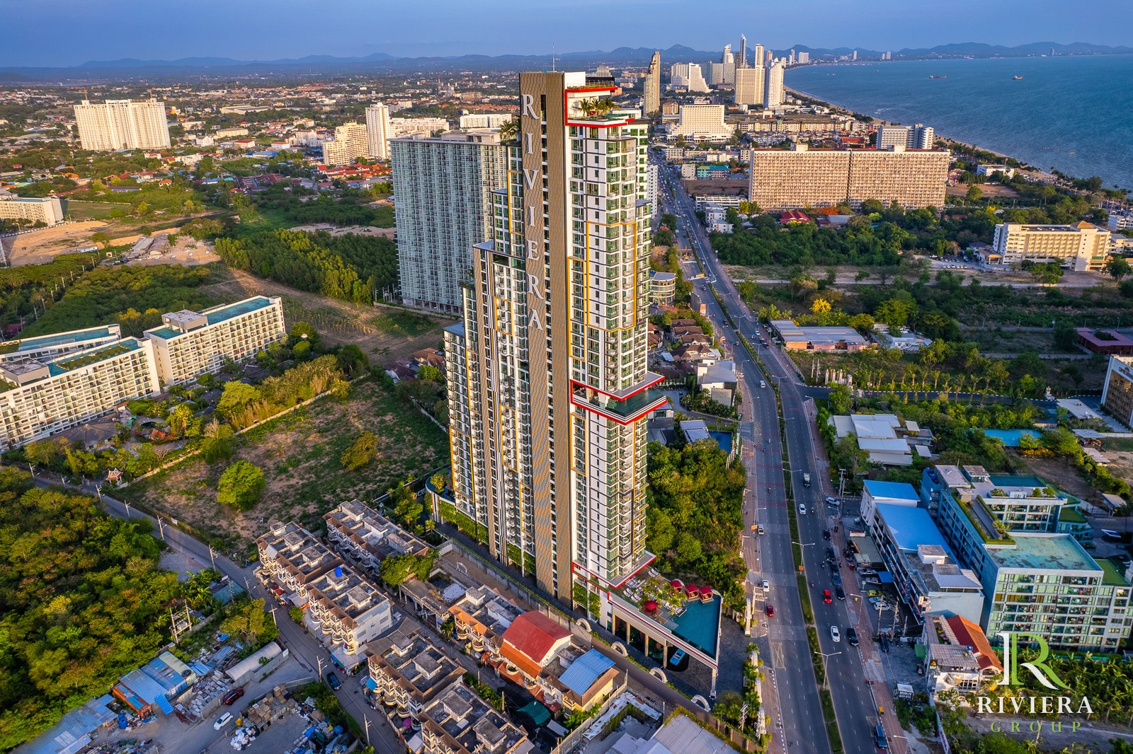 Aerial view of a tall residential tower near the coast with a city and sea in the background Riviera Group logo in the bottom right corner