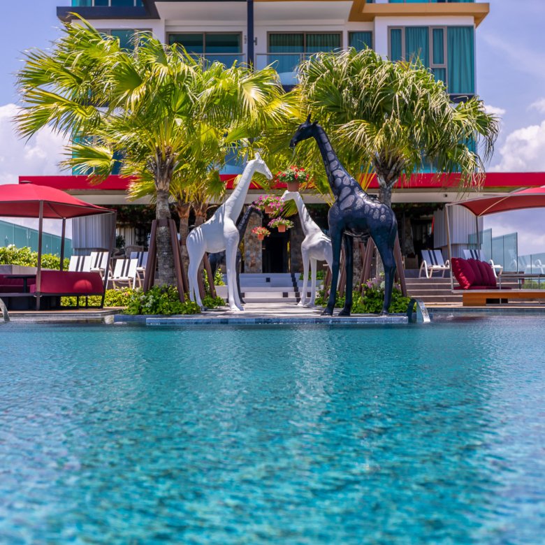 Poolside Riviera Group scene with three giraffe sculptures palm trees red shade canopies and modern buildings in the background