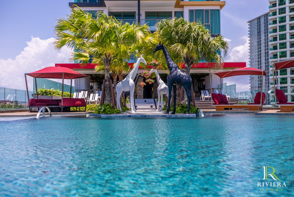 Poolside area with palm trees, red umbrellas, lounge seating, and tall decorative giraffe sculptures in front of modern buildings.
