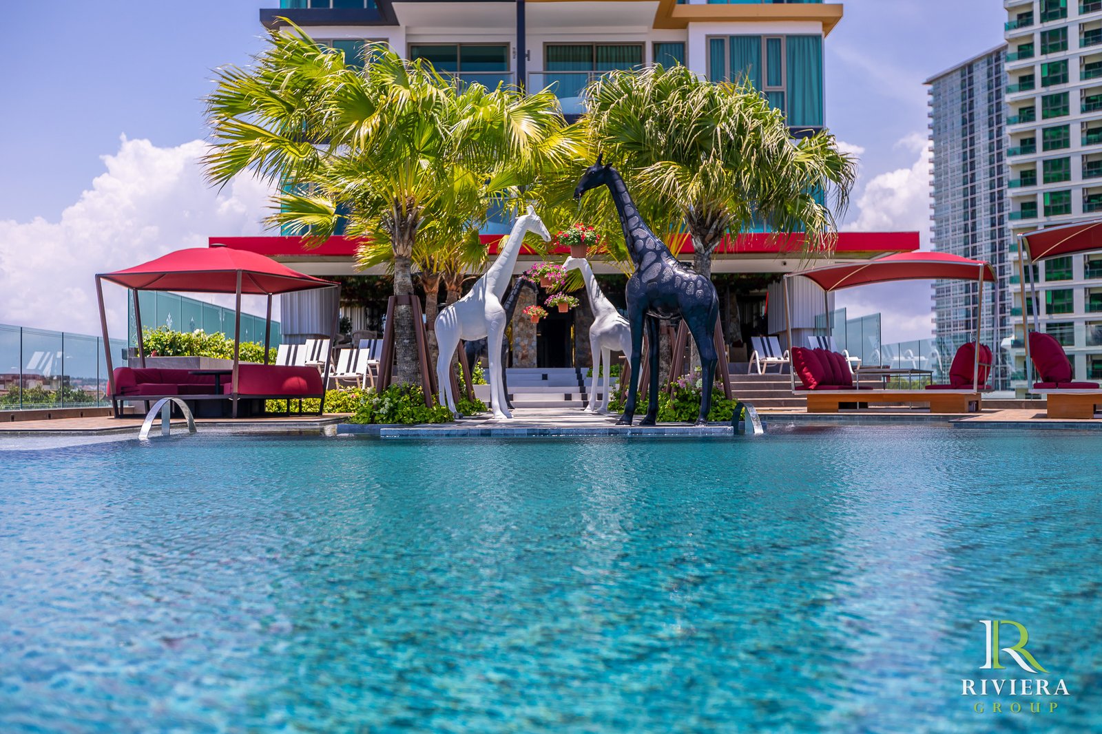 Luxurious hotel pool deck with palm trees, red canopies, and lounge seating plus decorative giraffe statues; Riviera Group branding visible.