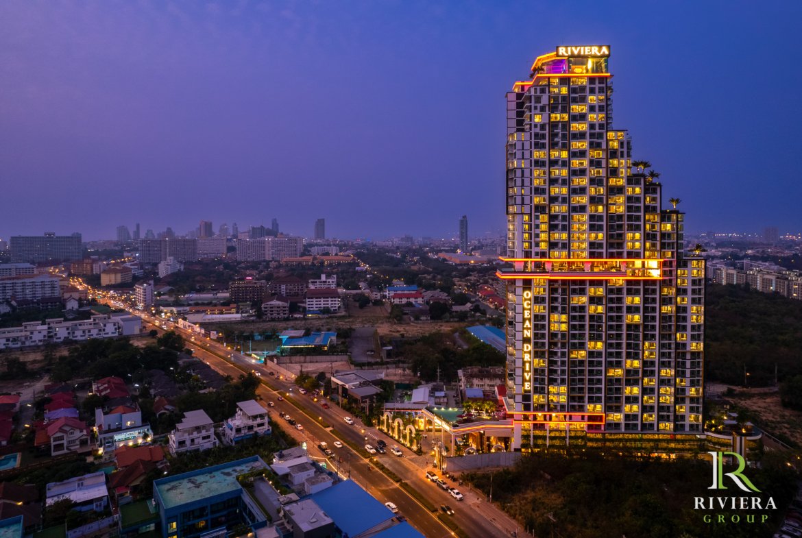 Night aerial view of a tall, neon-lit hotel with a 'RIVIERA' sign on top, overlooking a city at dusk.