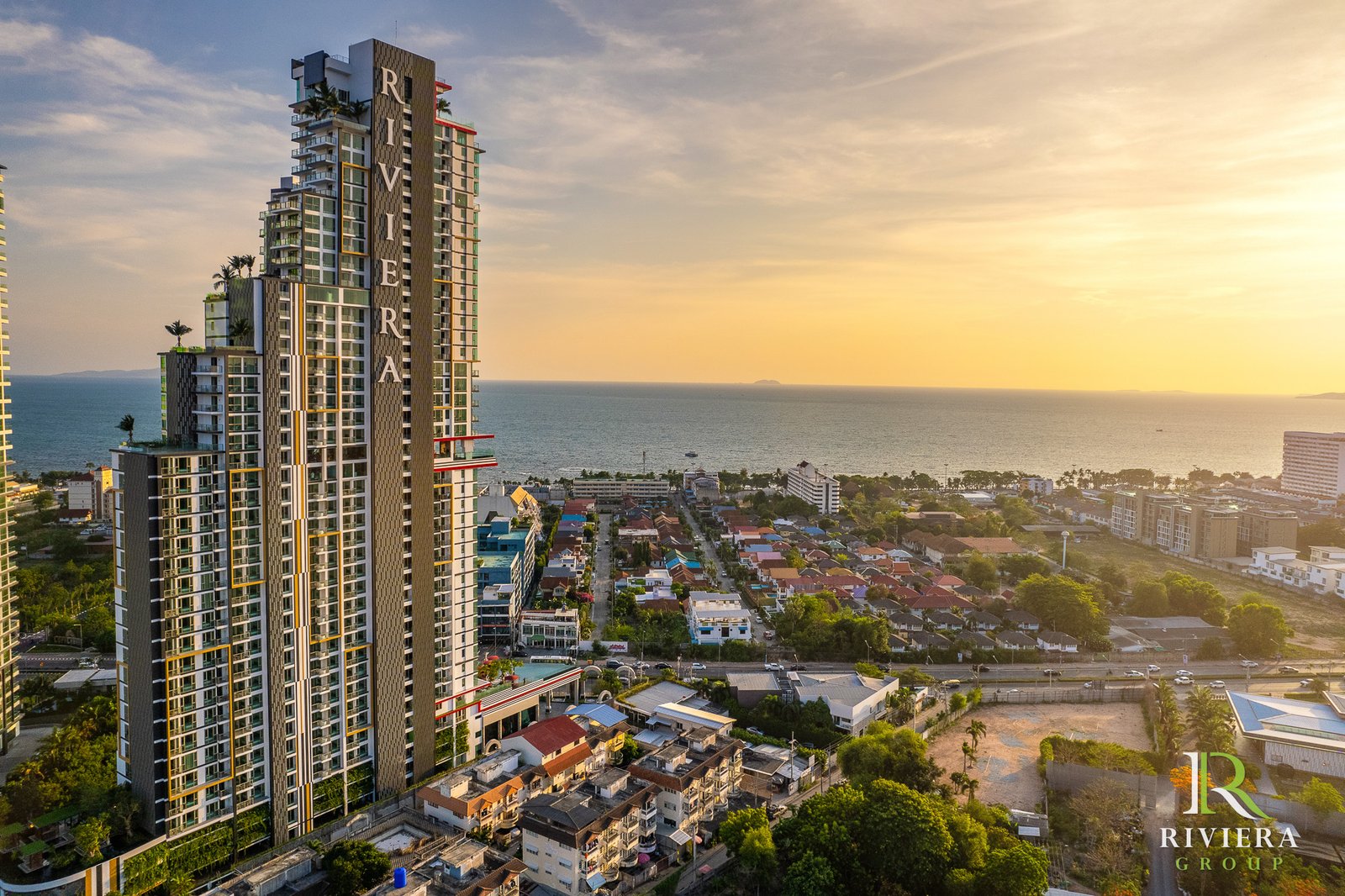 Tall modern beachfront condo tower with the Riviera logo in the lower-right, overlooking a coastal town at sunset.（informative）