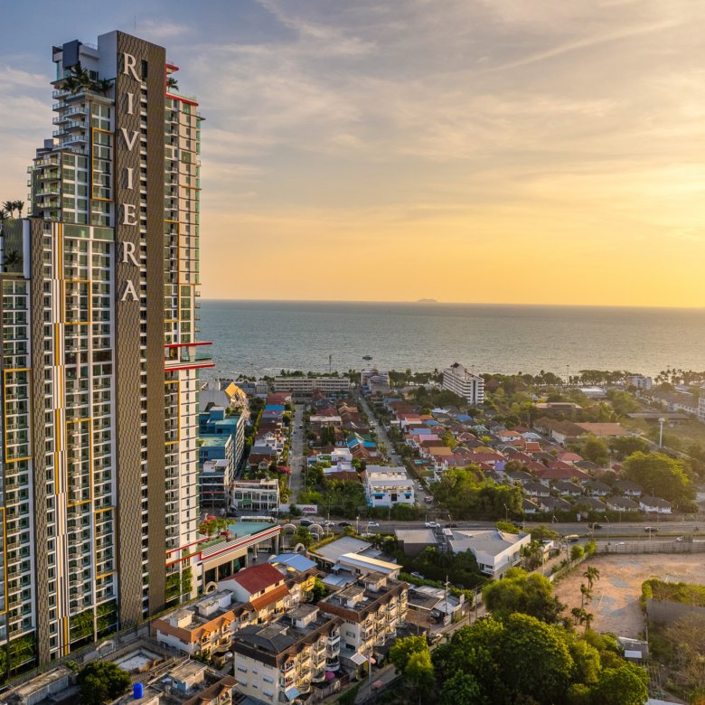 A tall modern residential tower by the sea at sunset with smaller houses and palm trees in the foreground and a calm ocean in the distance