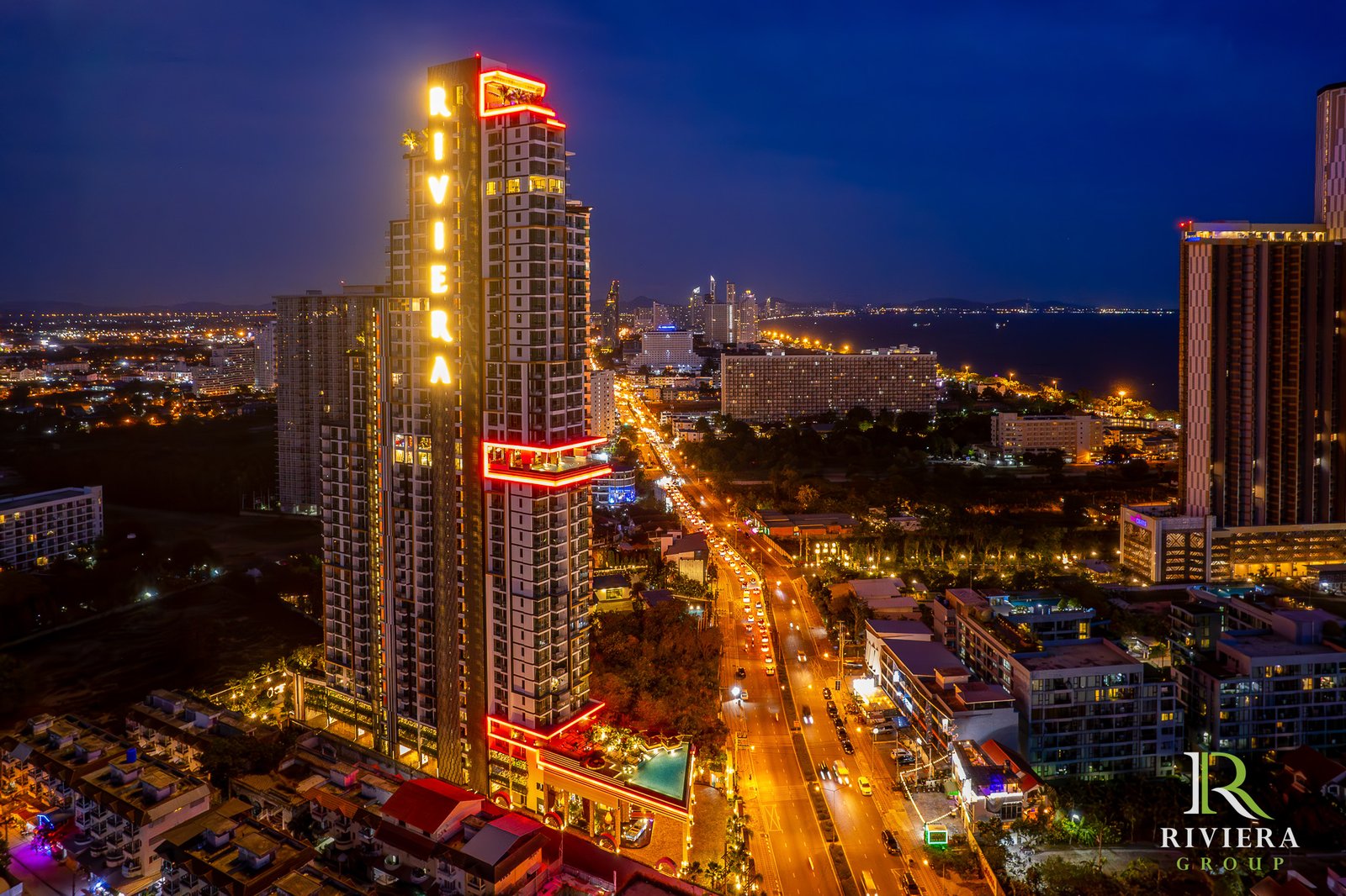 Riviera-Ocean-Drive-Pattaya 32 Nighttime city skyline with a tall, neon-lit high-rise prominently in the foreground and a busy street below; Riviera Group logo visible in the bottom-right corner.