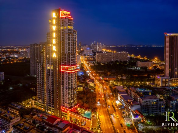 Aerial night view of a neon-lit high-rise building with red outlines and a vertical Riviera sign, overlooking a busy city street and coastline in the distance (RIVIERA GROUP logo bottom-right).