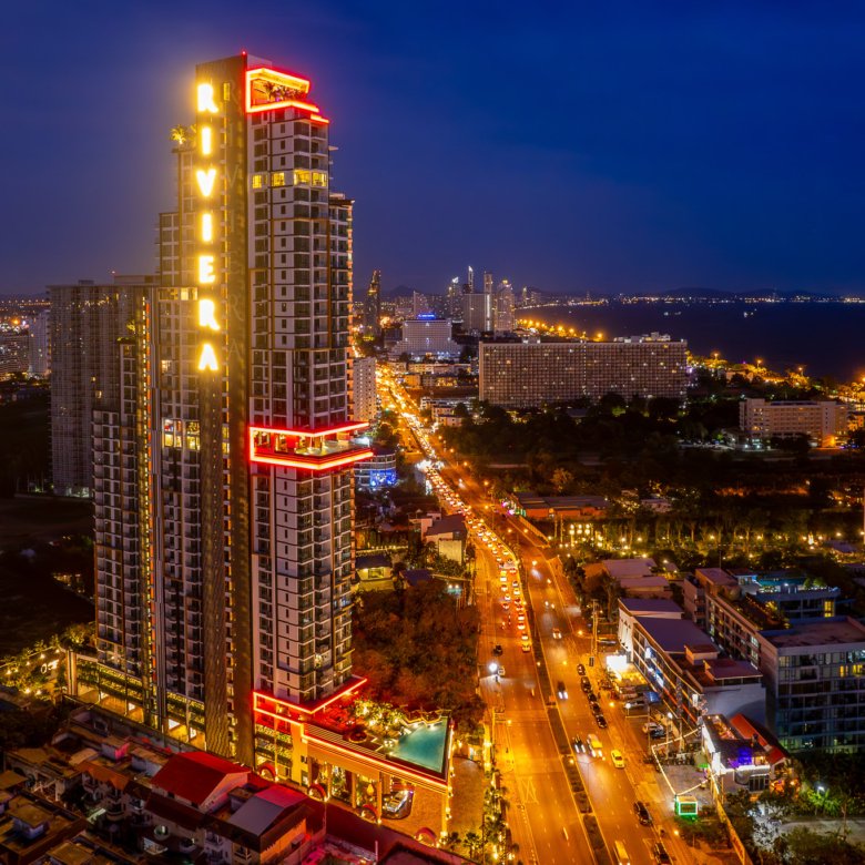 Aerial night view of a neon lit high rise building with red outlines and a vertical Riviera sign overlooking a busy city street and coastline in the distance RIVIERA GROUP logo bottom right