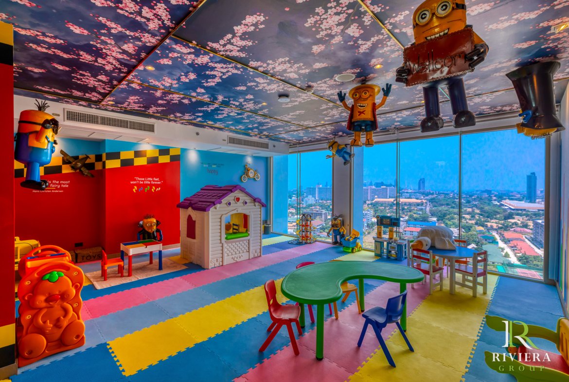Bright children's playroom with hanging Minion characters from the ceiling, colorful foam floor tiles, and a city view through large windows.