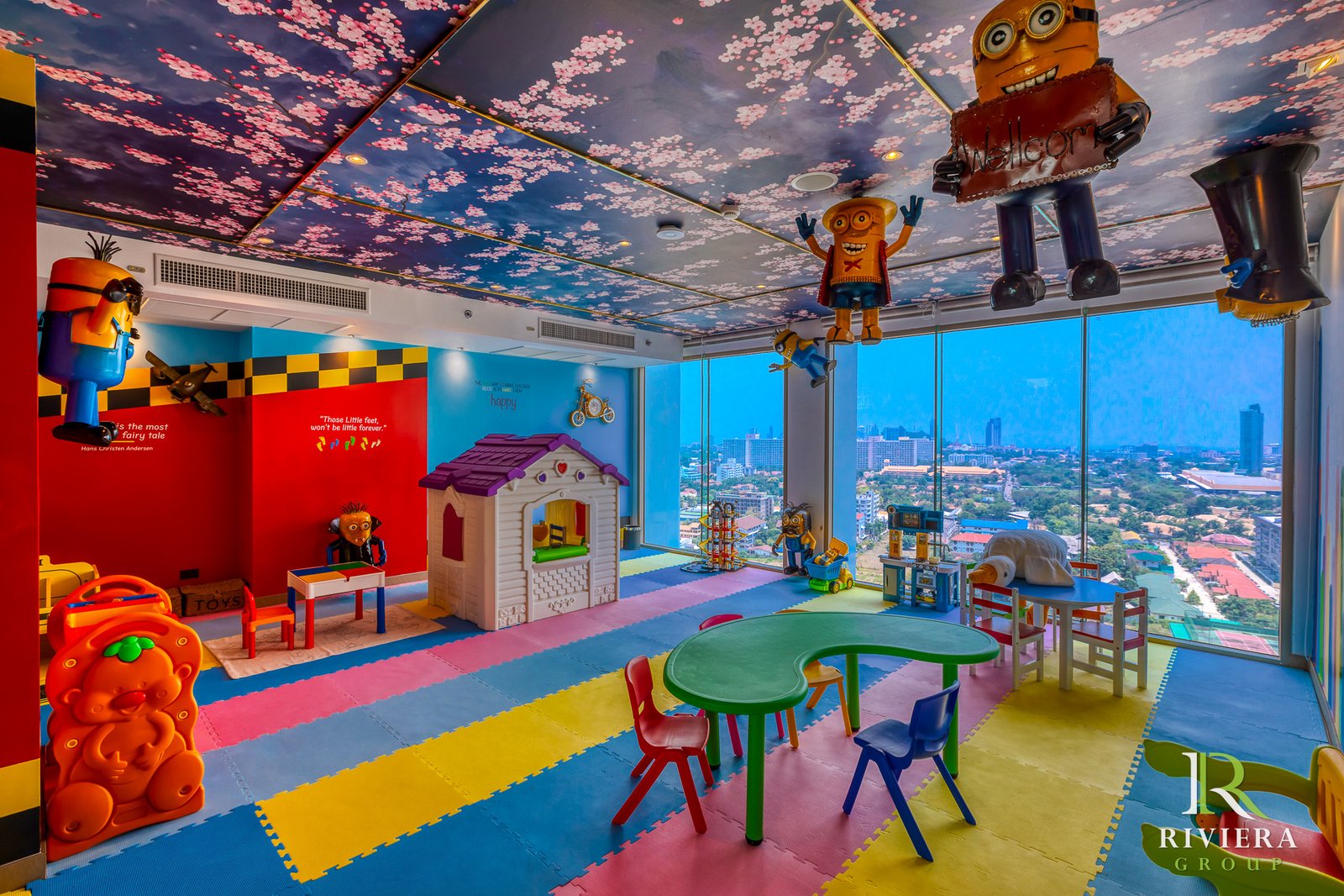 Riviera-Ocean-Drive-Pattaya 33 Colorful kids' playroom with Minion figures, toy house, and bright foam floor mats, overlooking a city skyline through large windows.