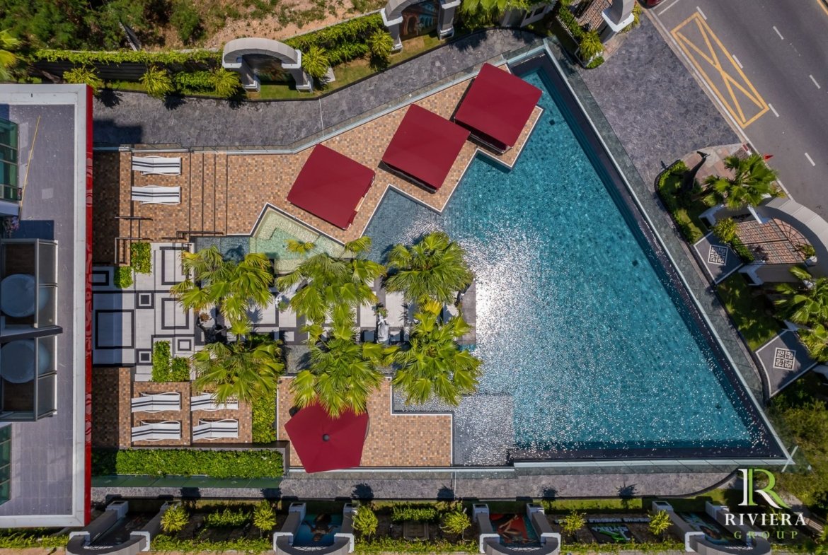 Aerial view of a resort pool area with red shade canopies, lounge chairs, palm trees, and a turquoise pool surrounded by tiled decking.