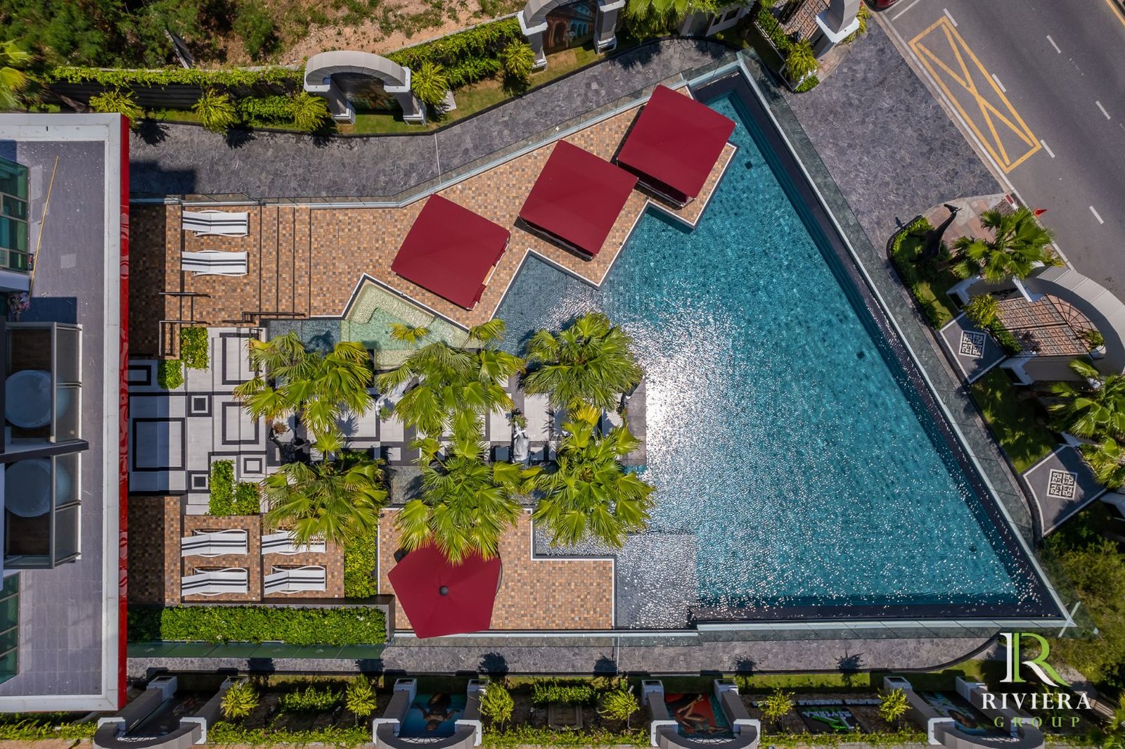 Riviera-Ocean-Drive-Pattaya 5 Aerial view of a resort pool area with red shade canopies, lounge chairs, palm trees, and a turquoise pool surrounded by tiled decking.