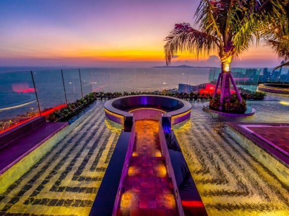 Sunset over the ocean seen from a luxury rooftop pool with glass railing and a palm tree, lit by colorful lights.
