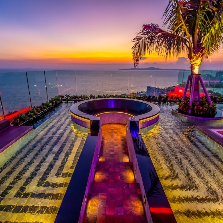 Sunset over the ocean seen from a luxury rooftop pool with glass railing and a palm tree lit by colorful lights