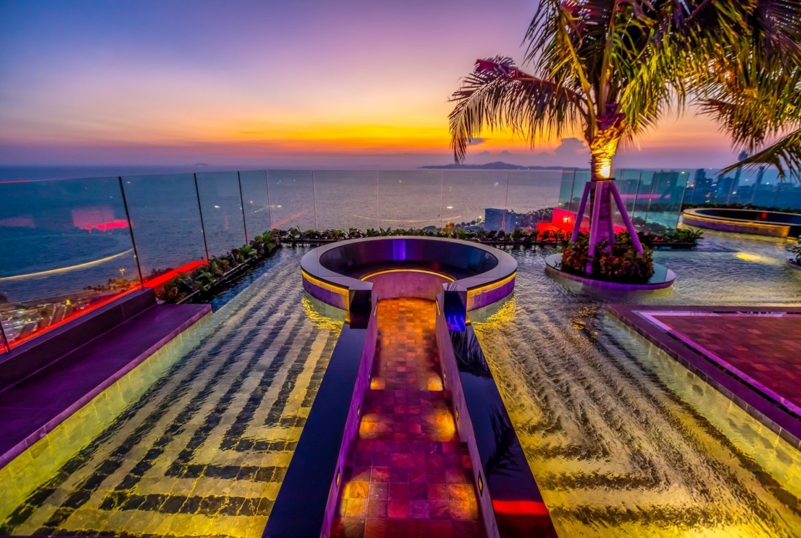 Rooftop pool at sunset with a circular seating area, palm tree, and glass railing overlooking the sea and distant lights.