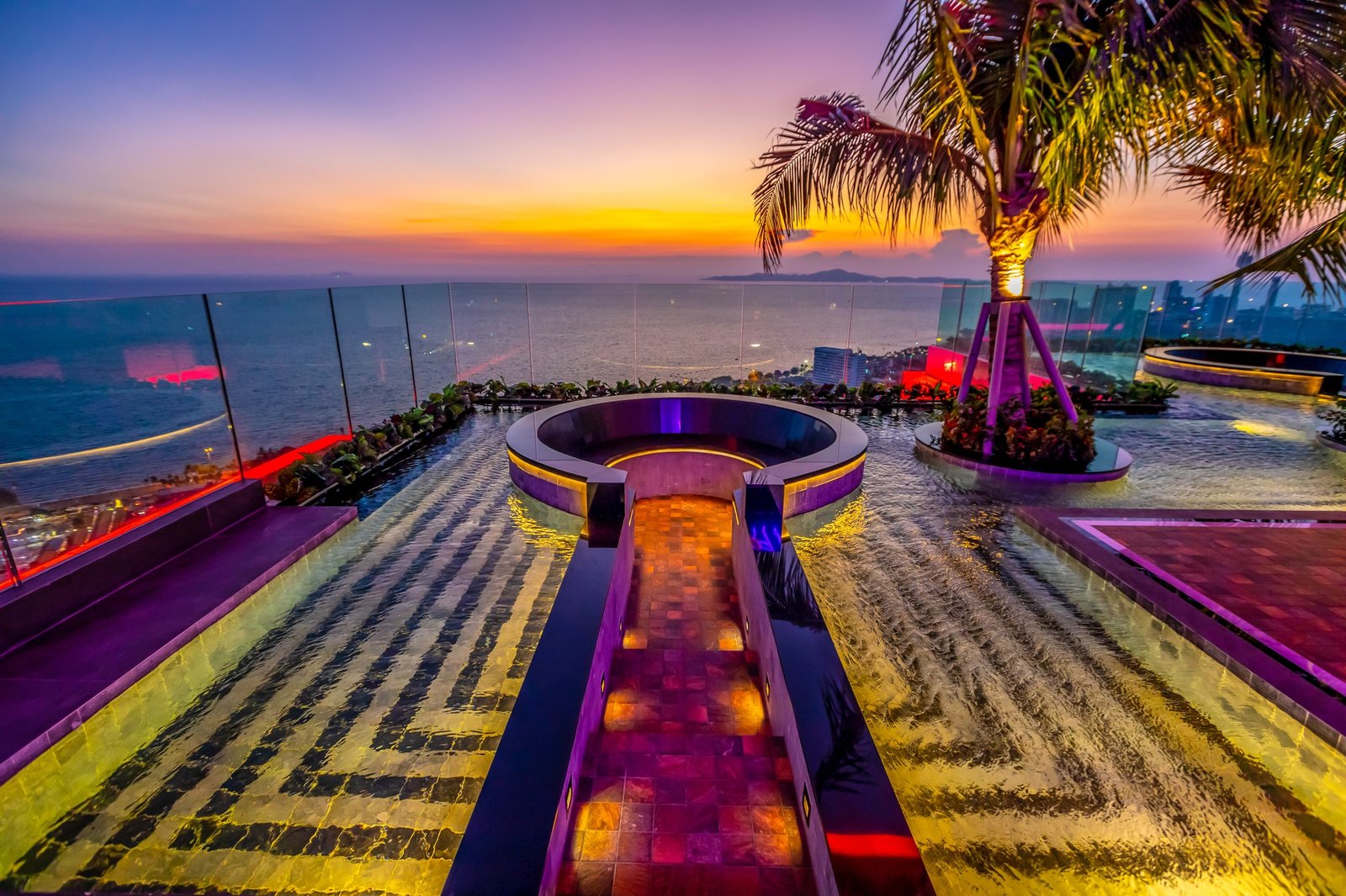 Riviera-Ocean-Drive-Pattaya 7 Rooftop infinity pool at dusk with glass railing, palm trees, and colorful lights over the ocean.